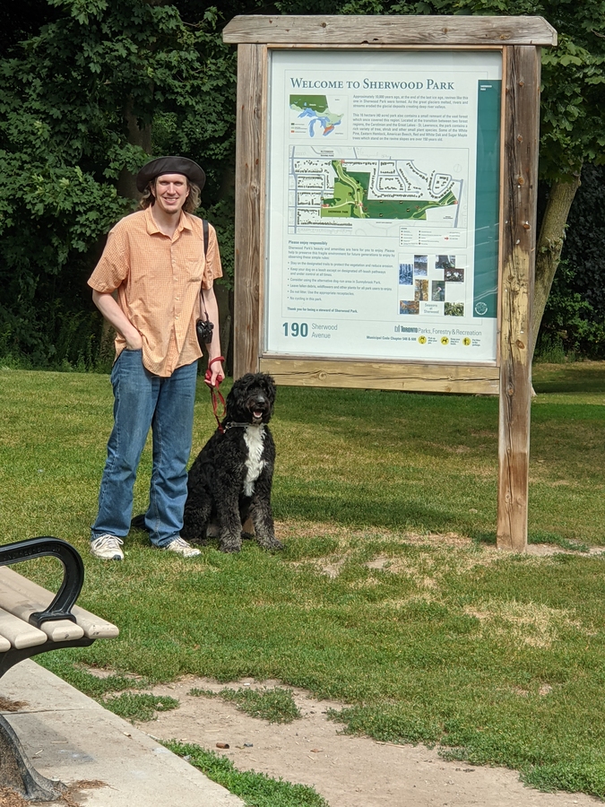 Sherwood Dog Park entrance sign with a sitting, fullsize, black and white Bernoodle and owner beside the sign.